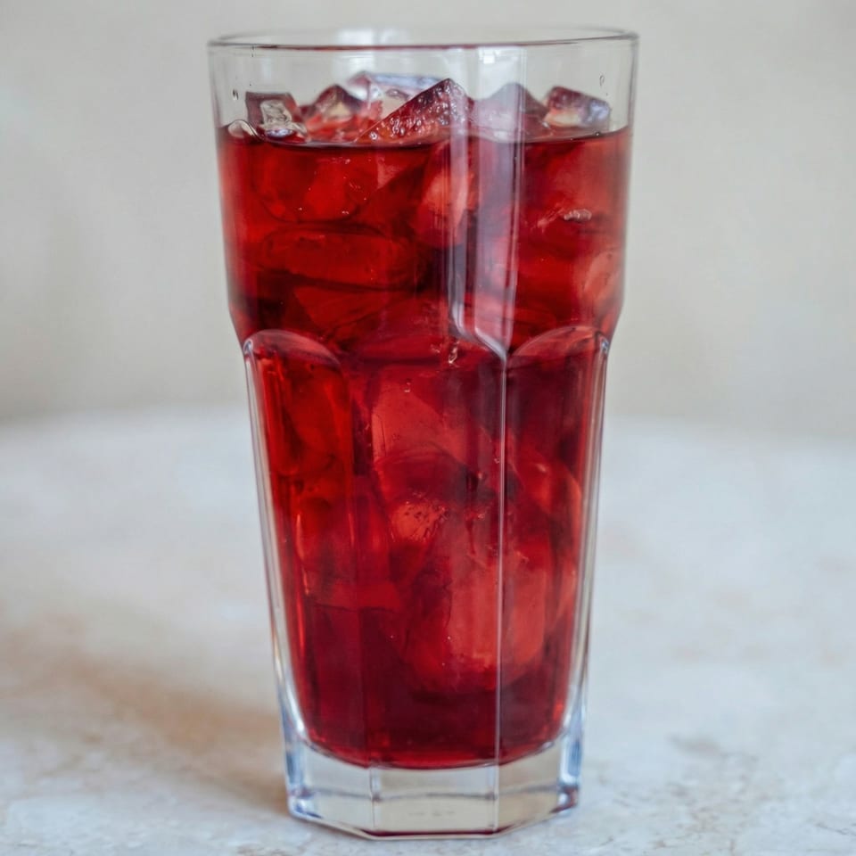 Tall glass filled with a vibrant red drink and ice cubes on a light-colored surface.