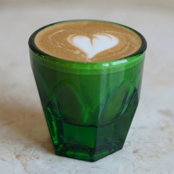 Latte with heart-shaped latte art in a faceted green glass on a marble surface.