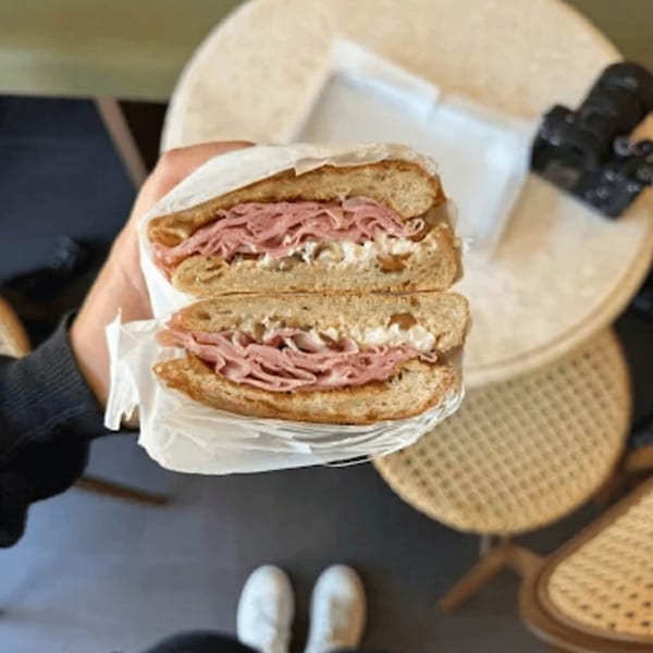 A person holds a sandwich with deli meat and cream cheese, cut in half, over a table.