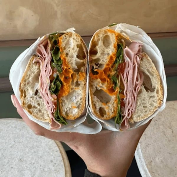 A hand holds a large sandwich, cut in half, revealing ham, greens, and orange sauce on crusty bread.