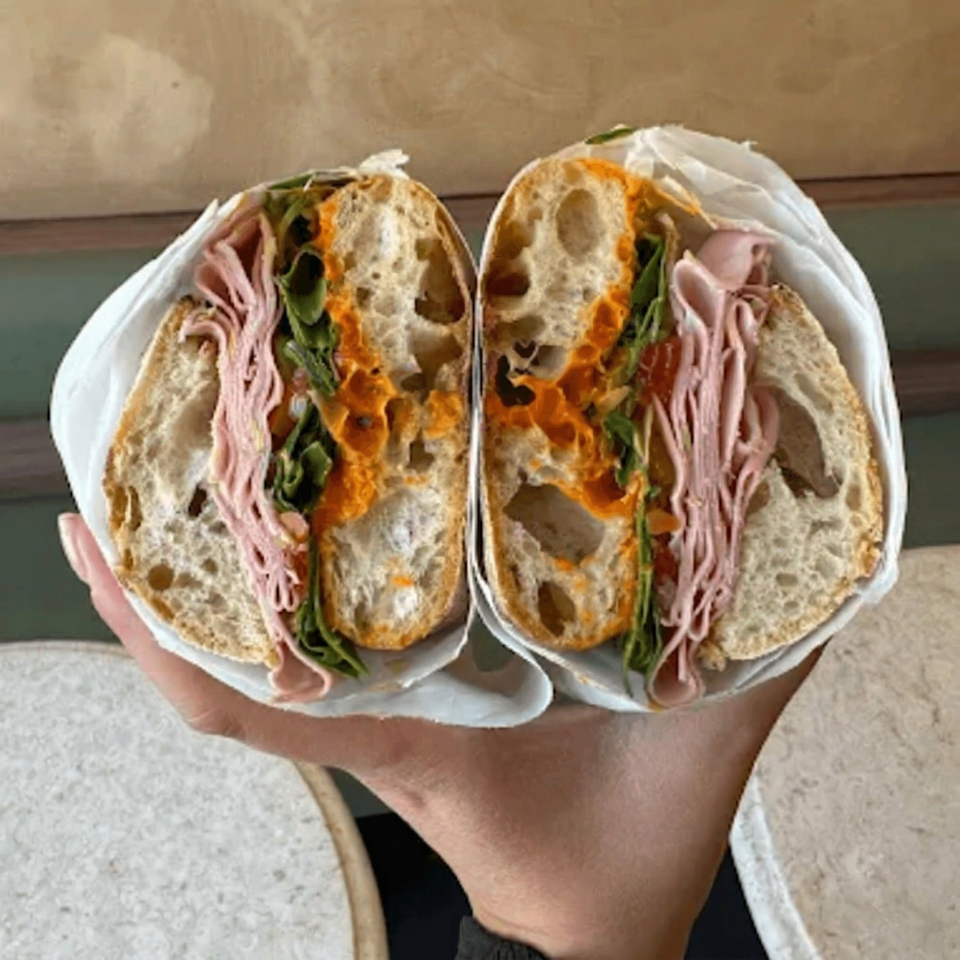 A hand holds a large sandwich, cut in half, revealing ham, greens, and orange sauce on crusty bread.